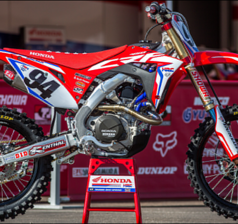 Red Honda dirt bike on a stand in a showroom/garage, displaying number 94 and multiple sponsor decals.