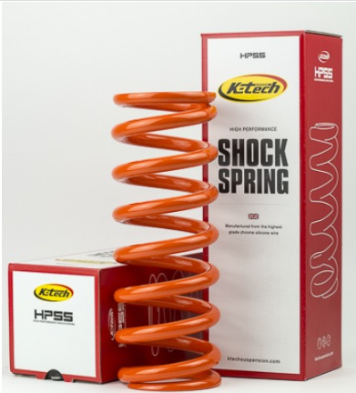 Bright orange coil spring shock absorber standing upright, with red-and-white K-Tech Shock Spring boxes in the background.