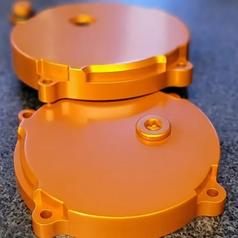 Two orange, circular anodized metal covers stacked to form a clamshell housing, with screw holes and a central fastener.