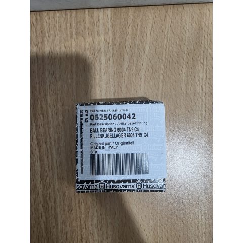 Small white box with a barcode and stamped text on a wooden surface; Part number 0625060042, describing a ball bearing 6004 TN9 C4, made in Italy, Husqvarna branding on tape around the box