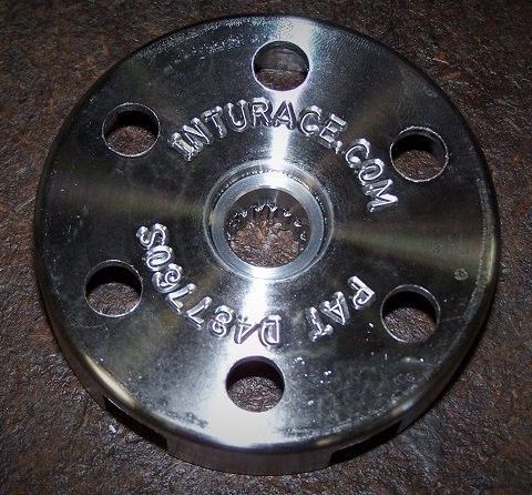 Shiny circular metal flange with a central bore and six evenly spaced outer holes, engraved with a website address on the surface, set on a dark, rusty surface.