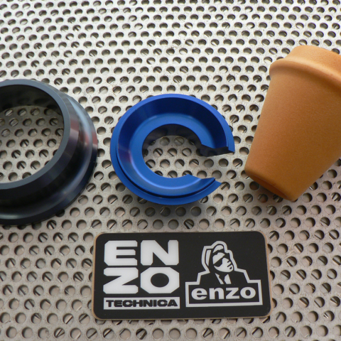 Disassembled blue C-shaped ring, black outer ring, and tan cup-shaped piece on a perforated metal surface, with an Enzo Technica label nearby.